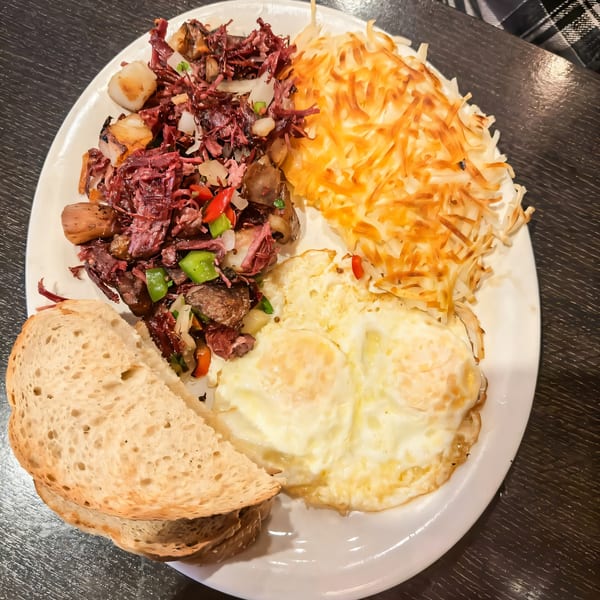 A breakfast plate with corned beef hash, crispy hash browns, two fried eggs, and toast.